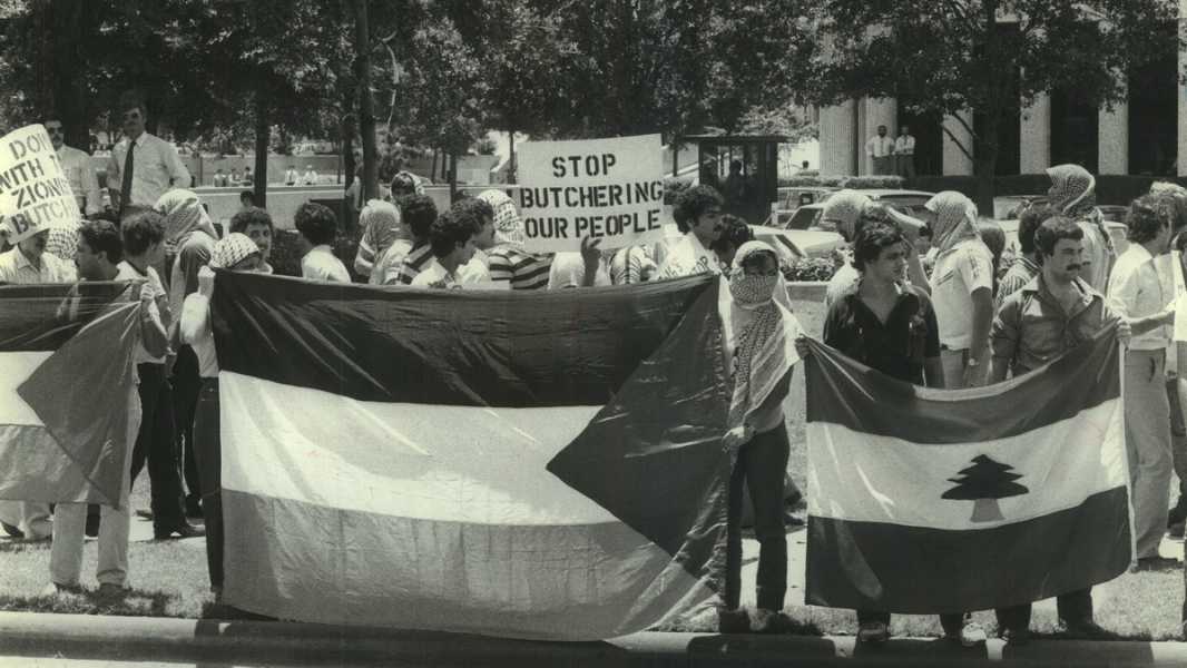 A photo of Palestinian students standing in front of a school building holding a Palestine Liberation Organization flag, left, and the Lebanese national flag, right. One student holds a sign that reads “Stop butchering our people.”