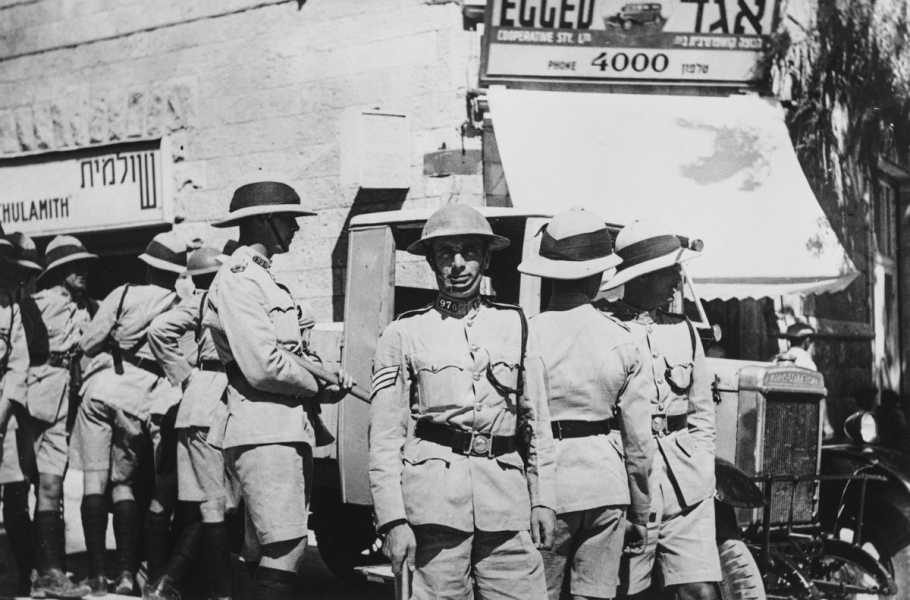 A black-and-white photograph of men in military uniforms and brimmed hats standing in front of shops bearing Hebrew signage.