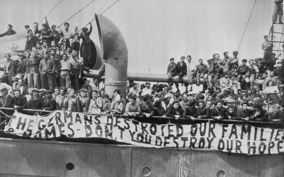 A black-and-white photo of a crowd of people aboard a ship’s deck. A large banner hanging over the side of the ship reads: “The germans destroyed our families ... don’t you destroy our hope.”