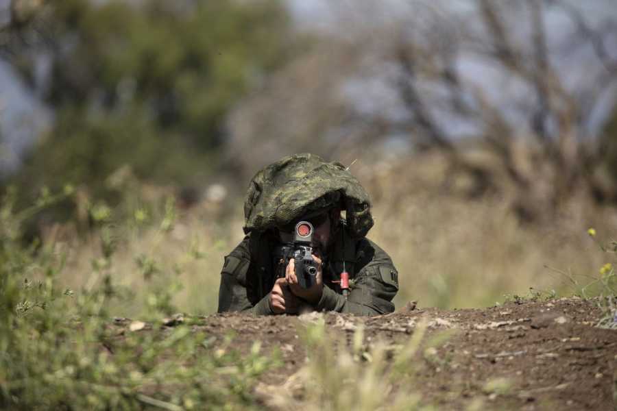 An Israeli soldier lying on the ground amid shrubs points a rifle directly at the camera.