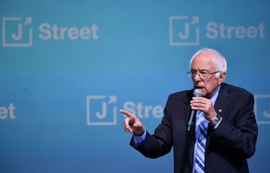 Bernie Sanders, in a dark suit and blue tie, speaks while holding a microphone, standing on a stage against a blue backdrop with “J Street” and the group’s logo.