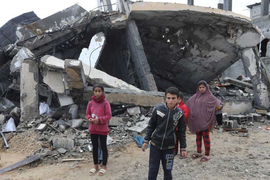 Several children stand in front of a collapsed concrete building, its structural supports sprawled across the ground. 