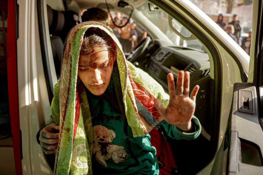 An injured girl with a scarf on her head holds up her hand as she steps out of the passenger seat of a van.