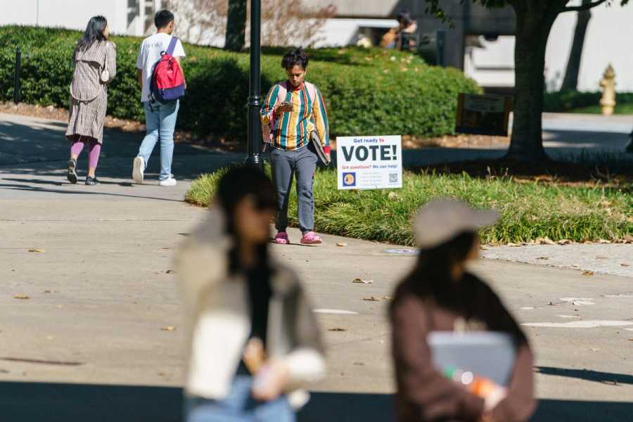 A sign reading “Vote!” is stuck into a grassy patch surrounded by cement sidewalks, where people walk in both directions past it, carrying backpacks and books.