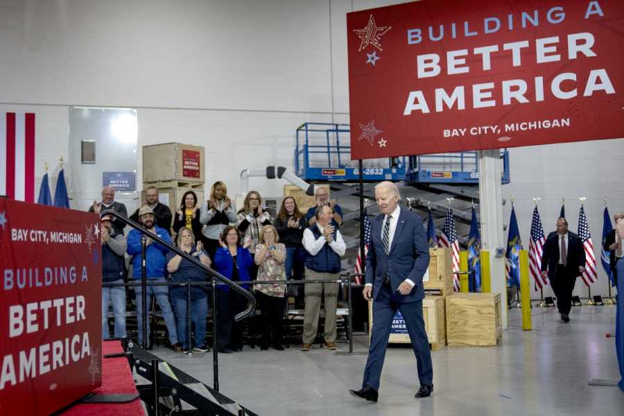 Joe Biden, pictured mid-stride, walks under a large red banner that reads: “Building a Better America.” A small group of people look on and clap.