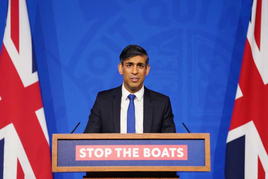Rishi Sunak, in a dark suit and blue tie, speaks from a podium reading “Stop the Boats” with the UK flag behind him. 