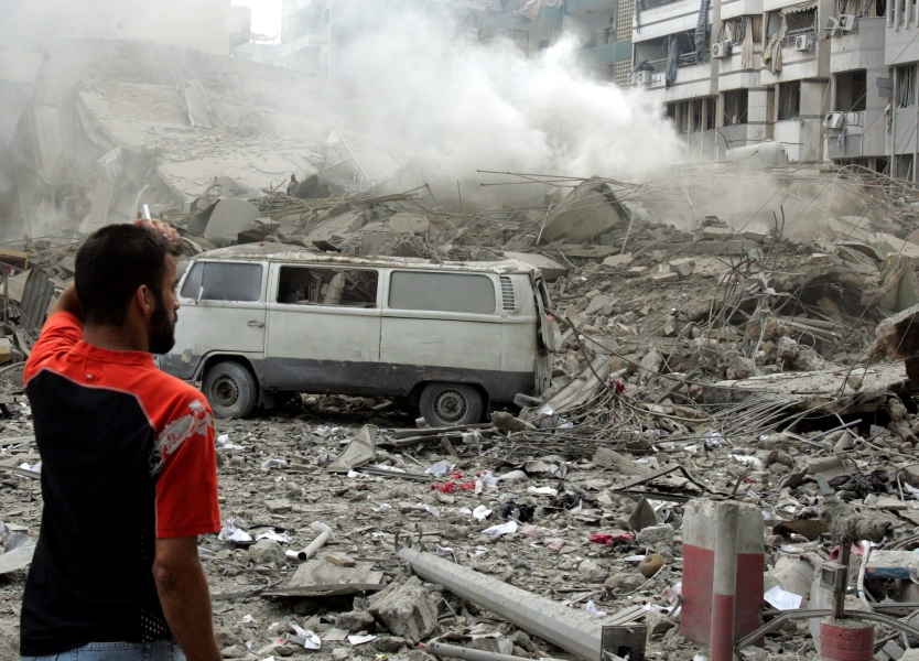 Smoke still rises from a flattened building that surrounds a silver van, its remaining windows thoroughly coated with grime. Debris covers the entire frame. The man, in an orange and black sports polo stands with his back mostly to us, his hand on his head as if in great sadness, desperation, or disbelief — perhaps all three.