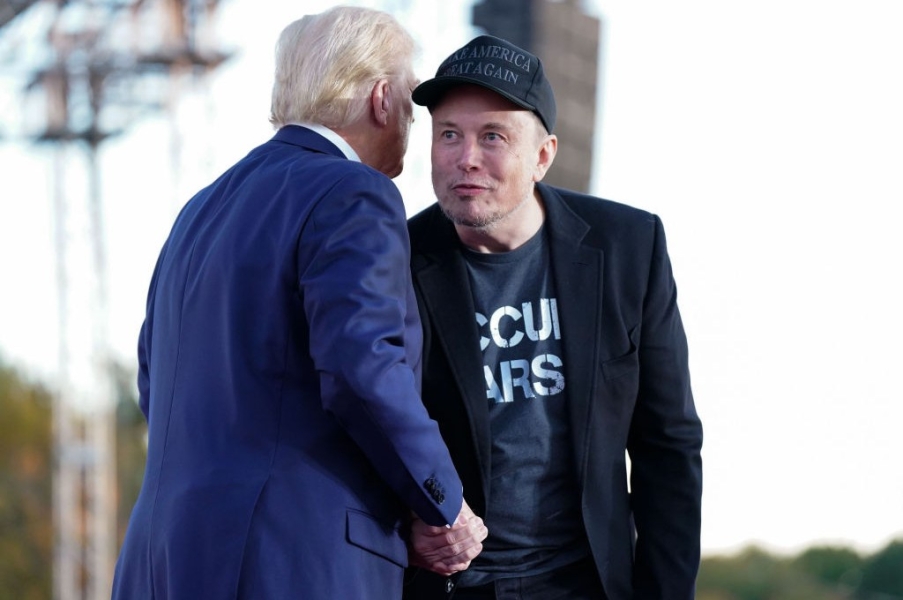 In a black Make America Great Again baseball cap and blazer, Elon Musk smiles as he shakes hands with Donald Trump, who is saying something in his ear.