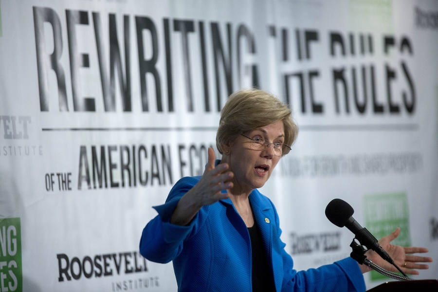 A photo of Elizabeth Warren speaking into a microphone in front of a sign which reads “Rewriting the rules”