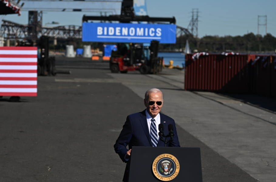 Biden speaks at a podium in an industrial outdoor space. On a piece of machinery behind him is a large sign reading “BIDENOMICS.”