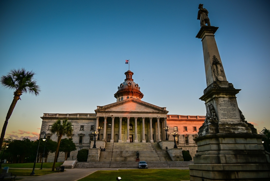 A grand building with a domed tower and columned front, flanked by palm trees and a stone monument.