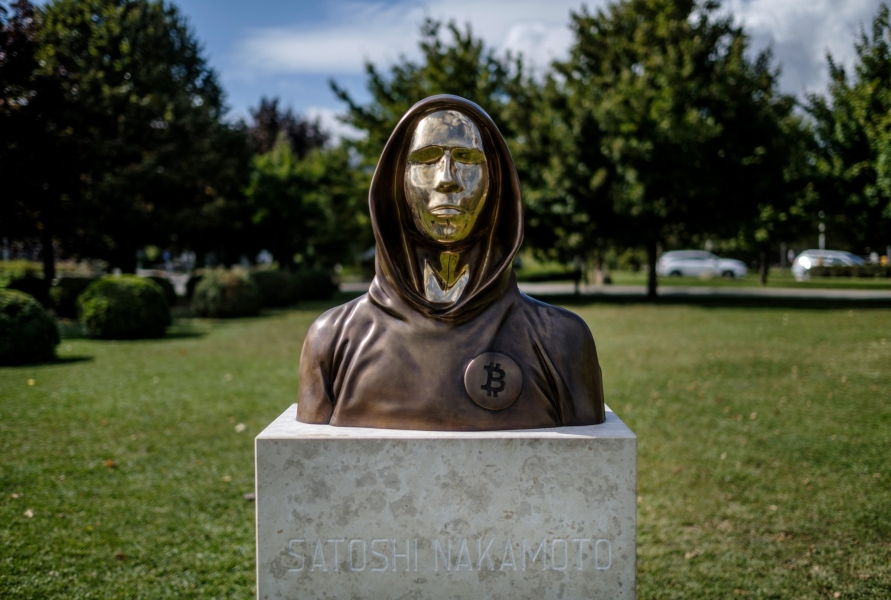 A gold statue with a bronze top and a hood is set atop a stone brick that reads Satoshi Nakamoto 