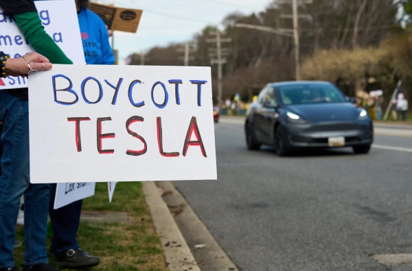 Protesters by the side of a road holding a sign that reads Boycott Tesla